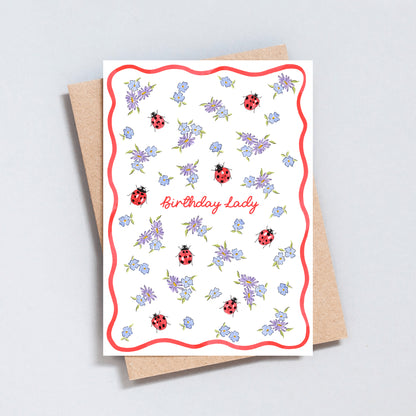 Illustrated Birthday Lady greeting card featuring hand-painted ladybirds, blue and lilac flowers and a red scalloped border.