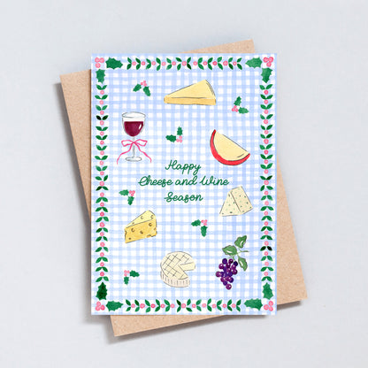 Illustrated Happy Cheese and Wine Season Christmas card featuring hand-painted cheeses, red wine, grapes and holly on a blue gingham background.