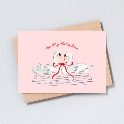 Romantic swan illustration Valentine’s card on a pink background with two swans tied together by a red ribbon.