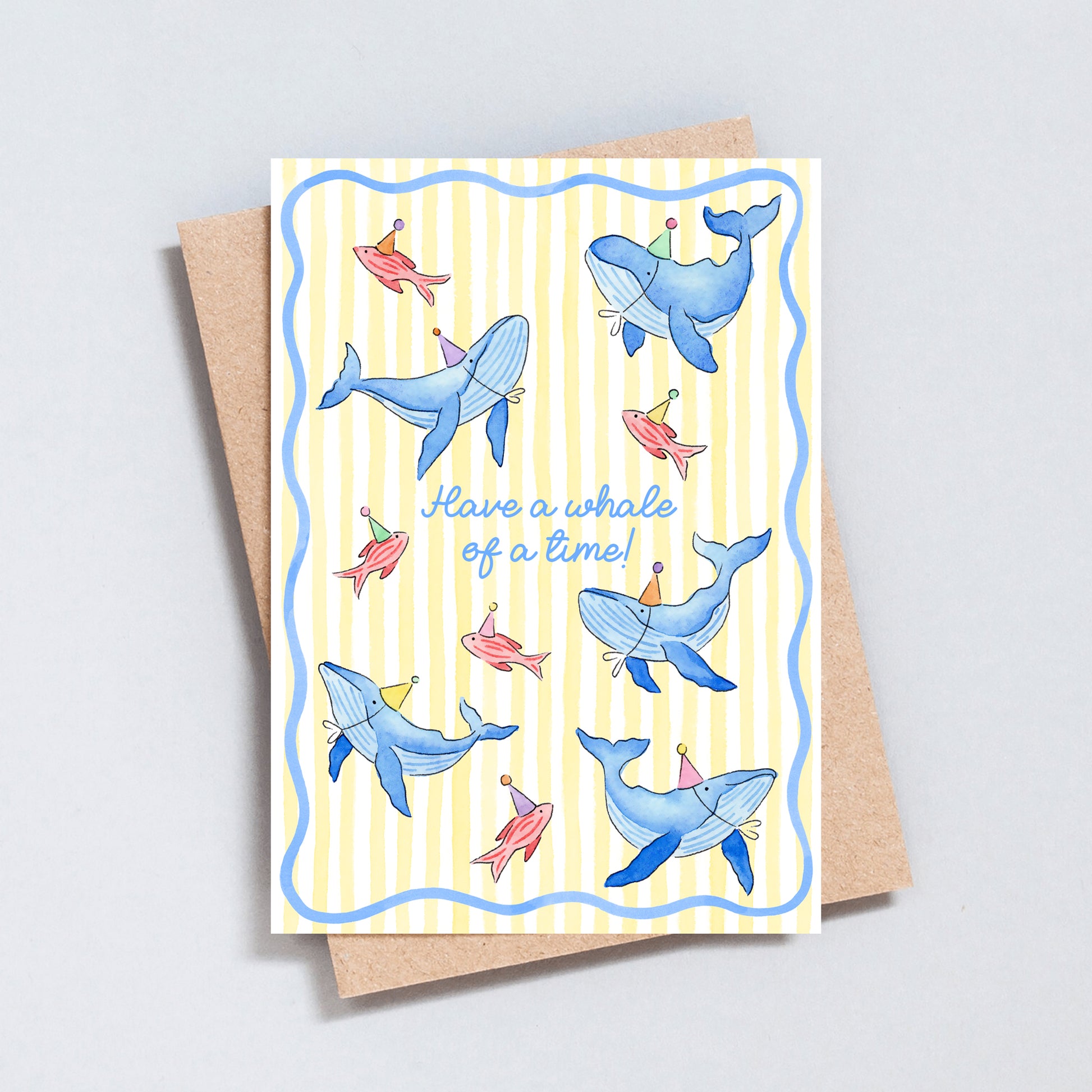Cute whale birthday card with illustrated blue whales and colourful fish wearing party hats on a pale yellow striped background, with the message “Have a whale of a time!”
